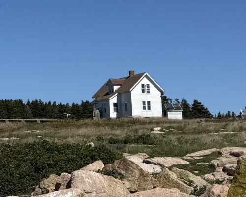 A white clapboard field station building with solar panels in a grassy, rocky field beneath a clear blue sky, with evergreen trees behind.