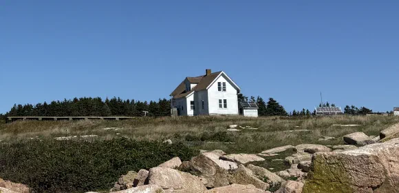 A white clapboard field station building with solar panels in a grassy, rocky field beneath a clear blue sky, with evergreen trees behind.