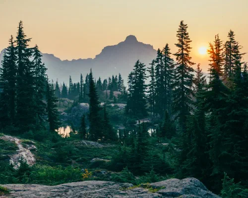 Sunset over a mountain wilderness with evergreen trees, rocky outcroppings, and a small reflective lake in the foreground