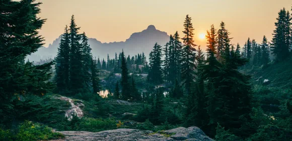 Sunset over a mountain wilderness with evergreen trees, rocky outcroppings, and a small reflective lake in the foreground